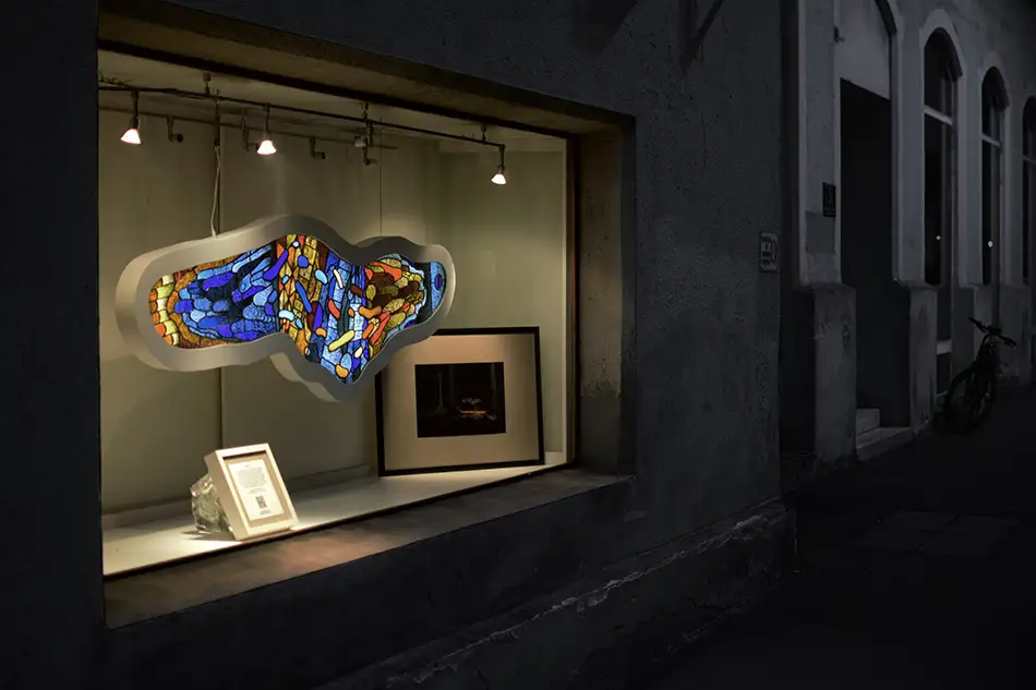 Exhibition view of a free-hanging and illuminated stained glass window with detailed painting which is named Amoeba and hangs in a forest