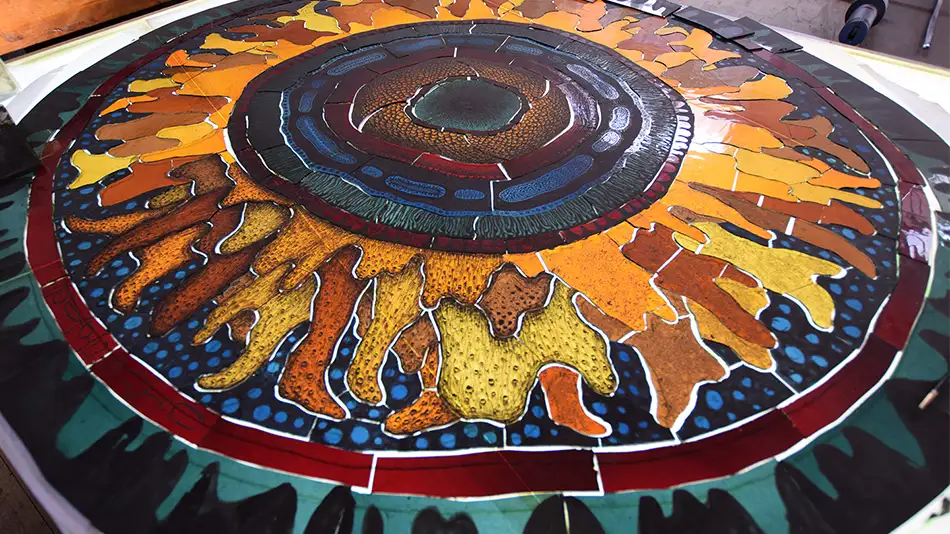 Exhibition view with people of a detailed painted stained glass window reminiscent of a gigantic fluorescent cell