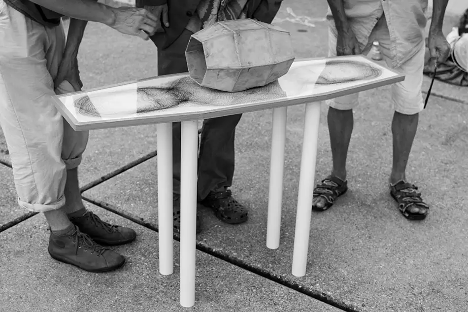 artwork made of concrete, mirror and digital print which is reminiscent of a table on which a geometric concrete element stands in the centre of which a mirror is fixed - with spectators